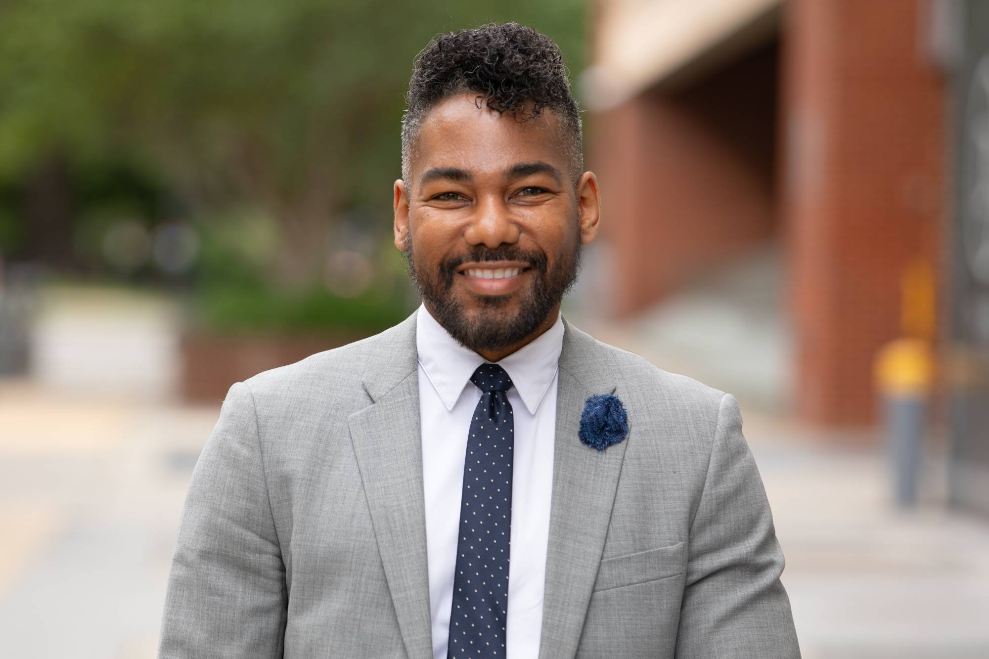 Smiling brown/black presenting man wearing a gray suit with a blue tie. Standing in a city.